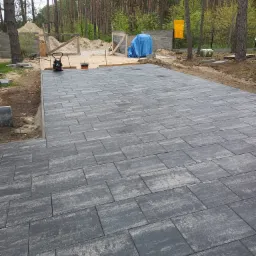 Ułożona szara kostka brukowa na posesji, widoczna ubijarka w tle, teren w trakcie prac wykończeniowych, drzewa w tle.