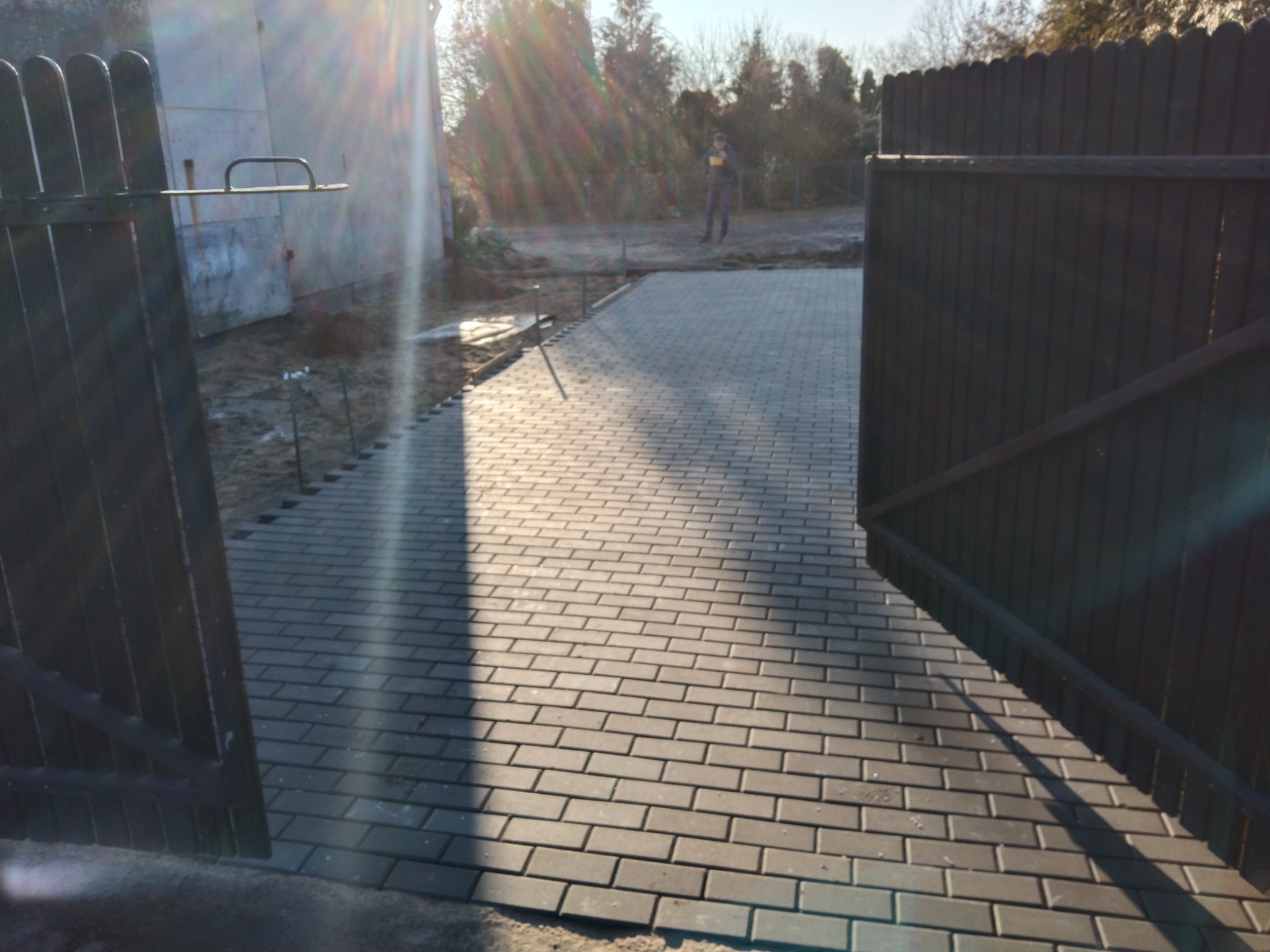 Nowo ułożona szara kostka brukowa w kształcie cegiełek, widoczna przez otwartą bramę. W tle osoba i budynek w trakcie budowy.