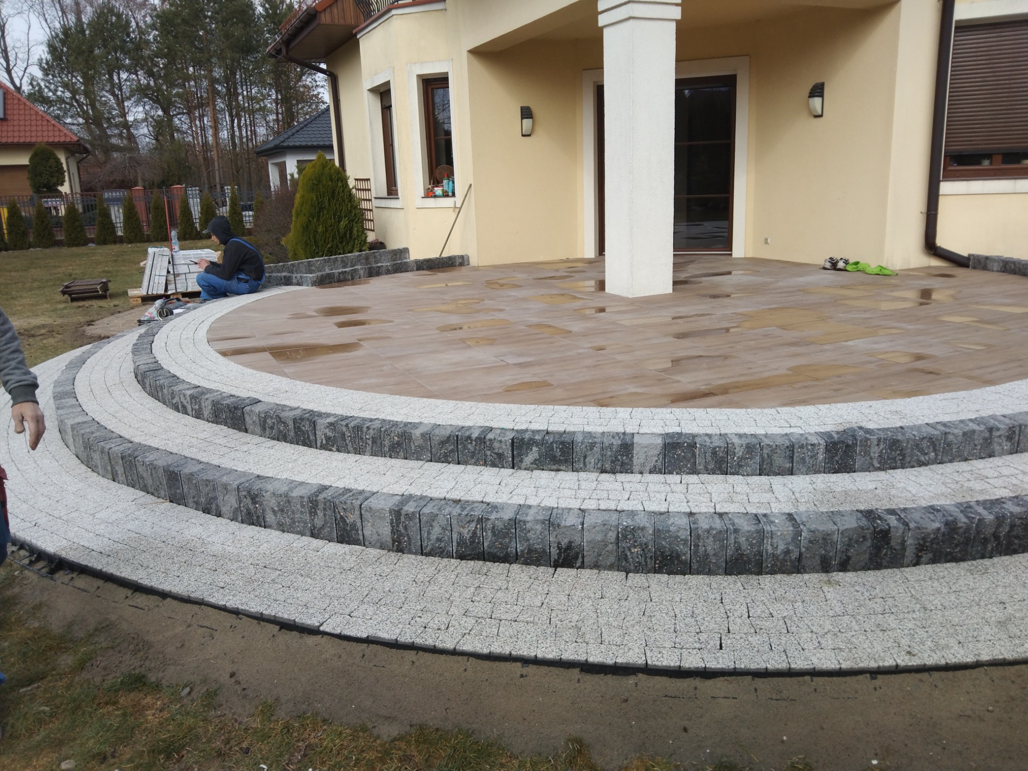 Półkoliste schody z kostki granitowej prowadzące na taras z płytek imitujących drewno, widoczne ślady wilgoci na płytkach, pracownik w oddali układa kostkę, dom jednorodzinny w tle.