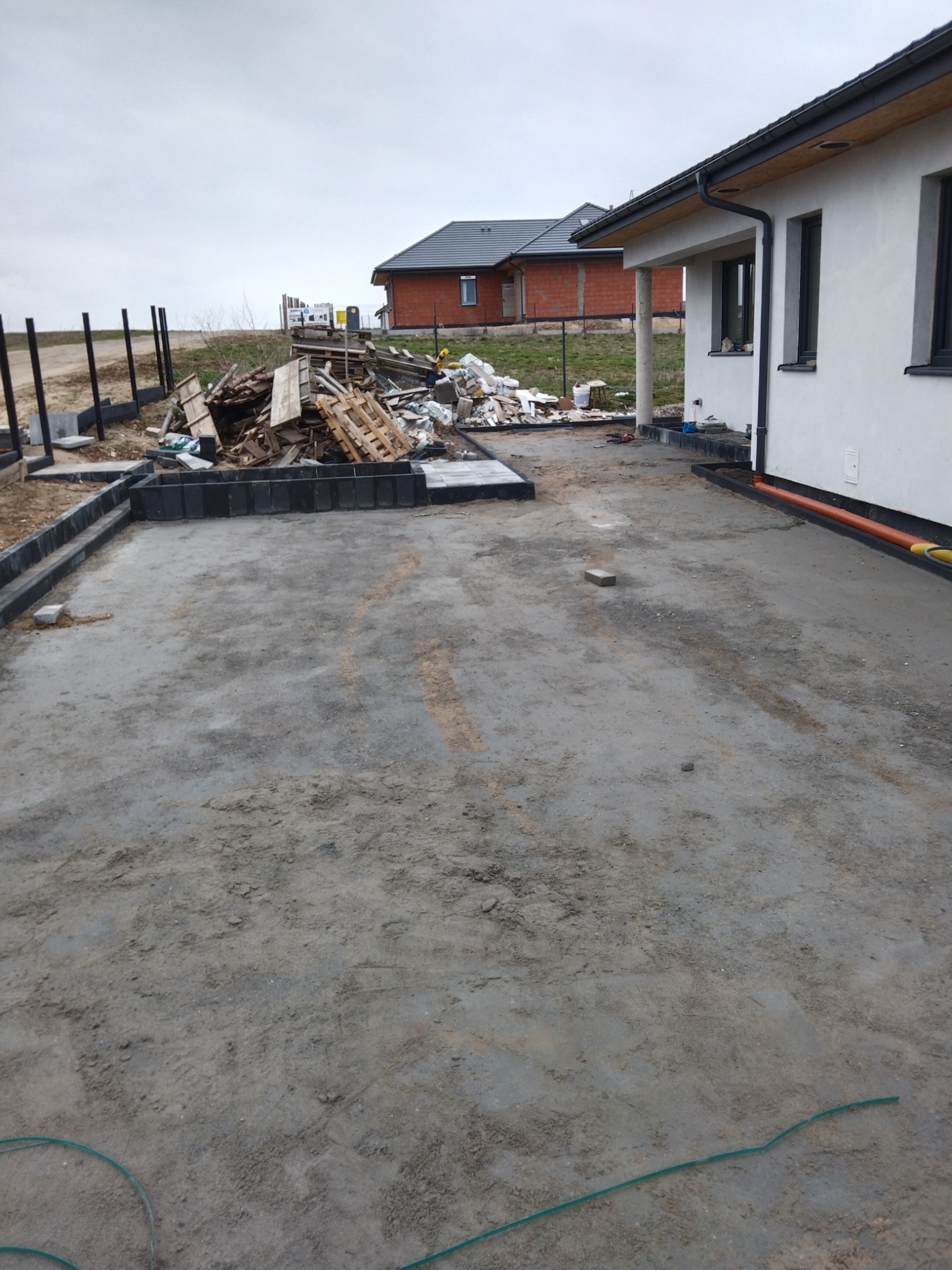 Przygotowany podkład z obrzeżami pod przyszłą nawierzchnię z kostki, widoczne palety i resztki materiałów budowlanych oraz rury drenażowe przy budynku.