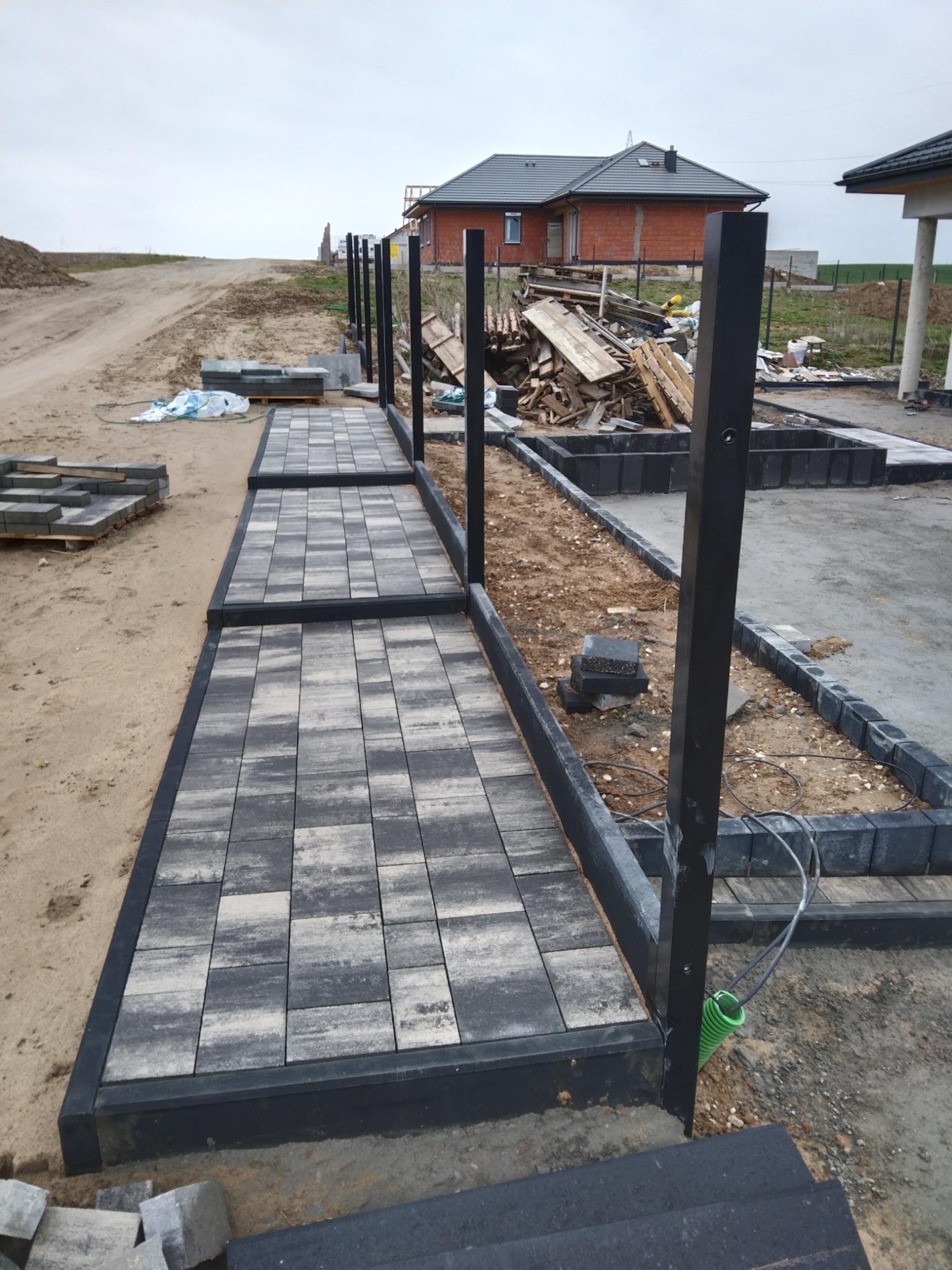 Układanie kostki brukowej wzdłuż ogrodzenia z czarnymi słupkami, widoczne przewody w zielonej osłonie, w tle budowa domu i sterta drewna.