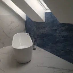 Nowoczesna łazienka na poddaszu z wolnostojącą białą wanną, marmurowymi płytkami podłogowymi i niebieskimi płytkami ściennymi imitującymi kamień, oświetlona światłem wpadającym przez okno dachowe.