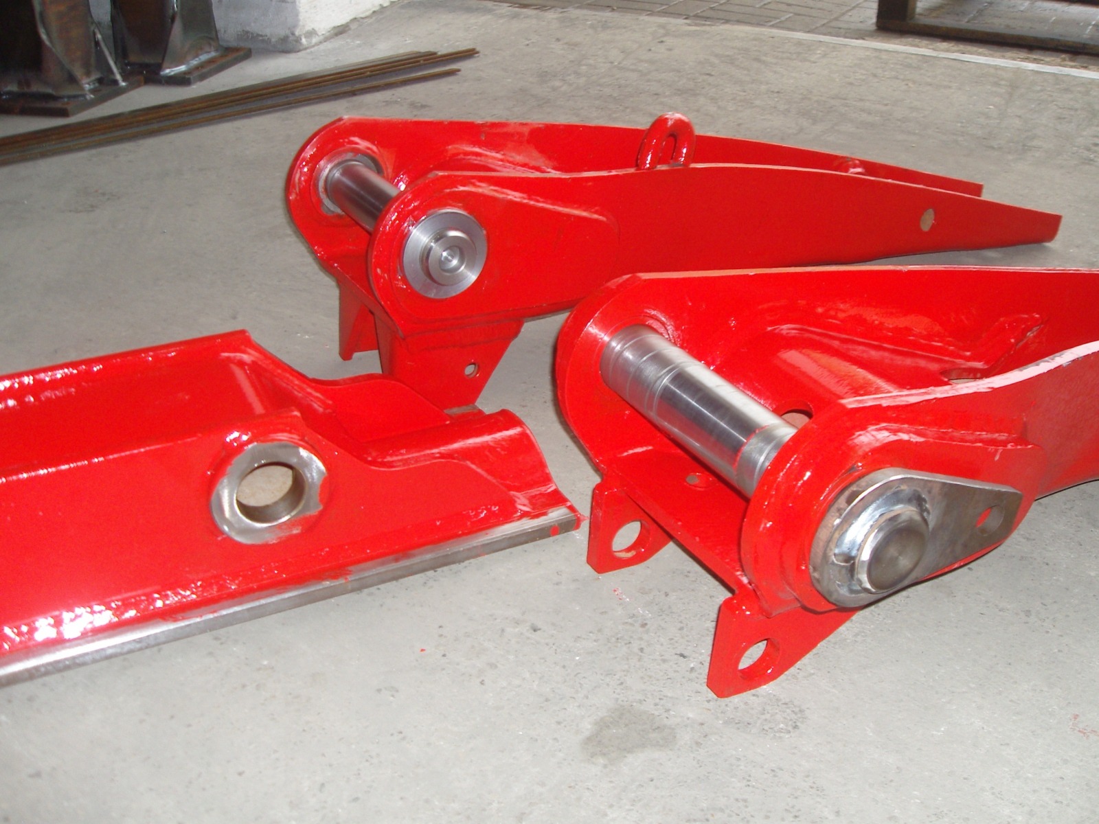Czerwone, pomalowane elementy konstrukcyjne z metalowymi tulejami, prawdopodobnie ramiona koparki, leżące na betonowej posadzce.