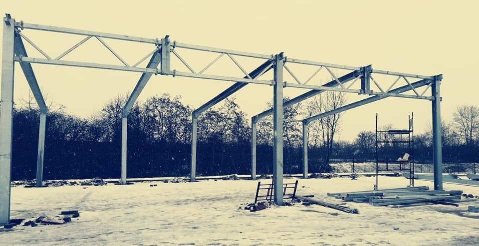 Stalowa konstrukcja hali w trakcie montażu, widoczne elementy szkieletu i rusztowanie na zasypanym śniegiem placu budowy.