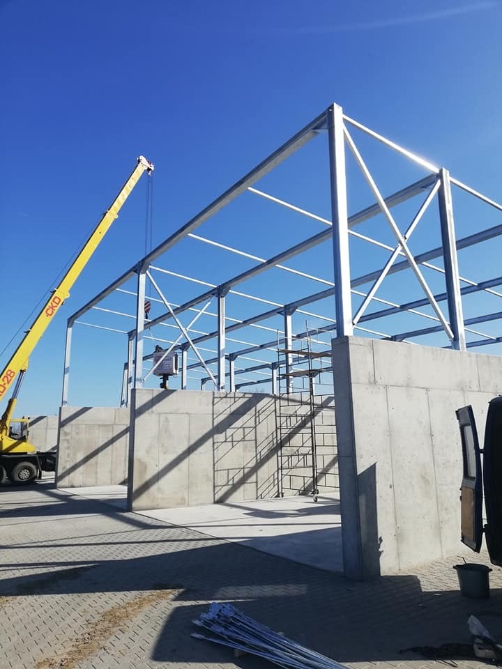 Wznoszenie konstrukcji stalowej hali: widok na szkielet z profili stalowych, betonowe ściany, żółty dźwig z wysięgnikiem, drabina rusztowaniowa, jasne niebo.