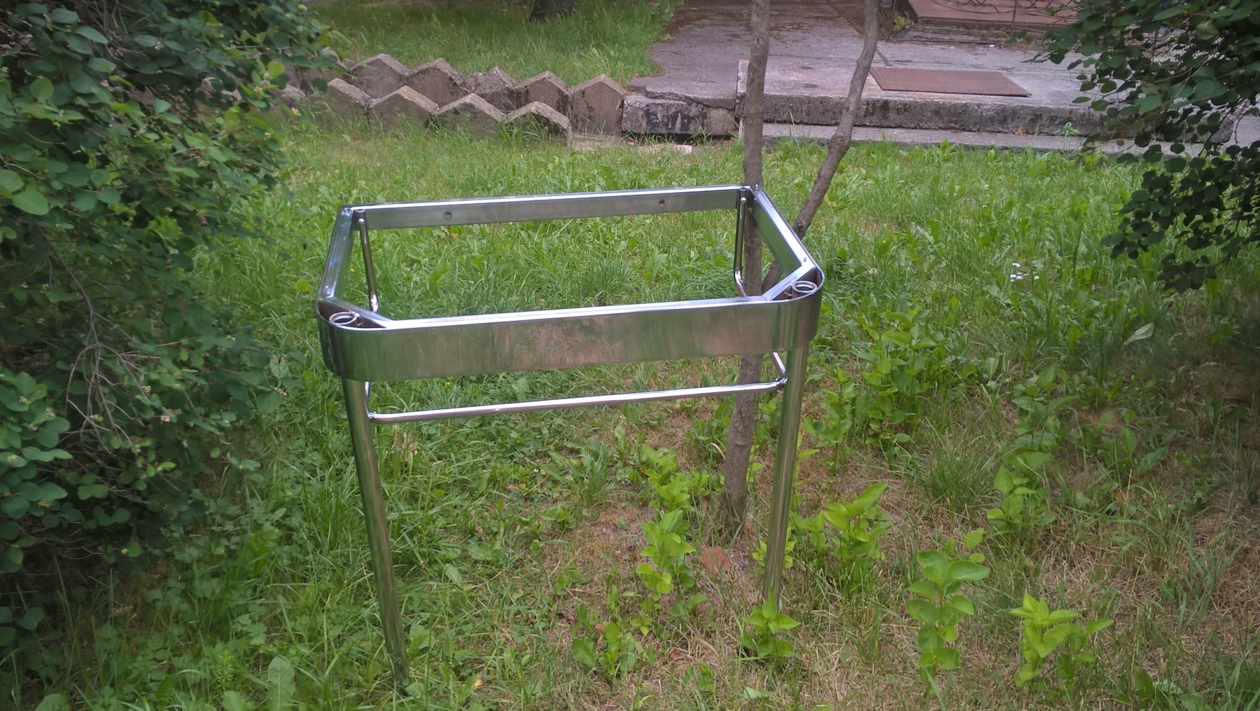 Chromowana, metalowa konstrukcja na czterech nogach, stojąca na trawie obok cienkiego drzewa. Widoczne spawane łączenia i polerowana powierzchnia.