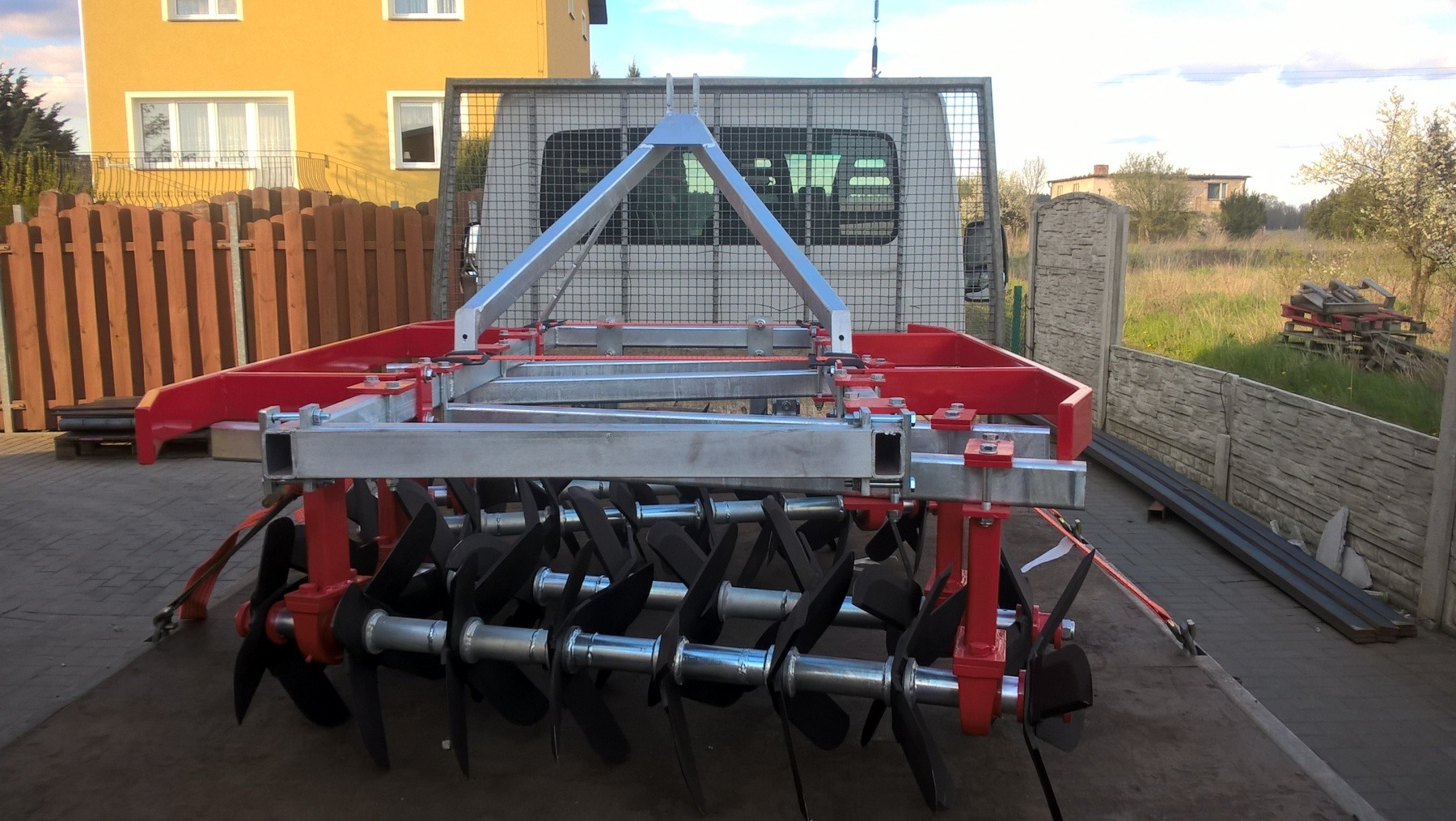 Czerwono-srebrna brona talerzowa, częściowo załadowana na przyczepę samochodu dostawczego, widoczna na tle zabudowań i ogrodzenia.