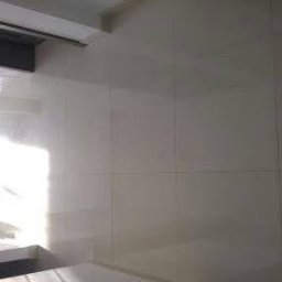 Jacek Żuchowski - Błyszczące, białe płytki ceramiczne na ścianie łazienki, z odbiciem światła w fugach i widocznym fragmentem sufitu oraz podłogi, z perspektywy pod kątem.