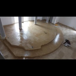 Jacek Żuchowski - Układanie beżowych, kwadratowych płytek na podłodze w pomieszczeniu z kolumnami; pracownik nanosi zaprawę na krawędź podestu.