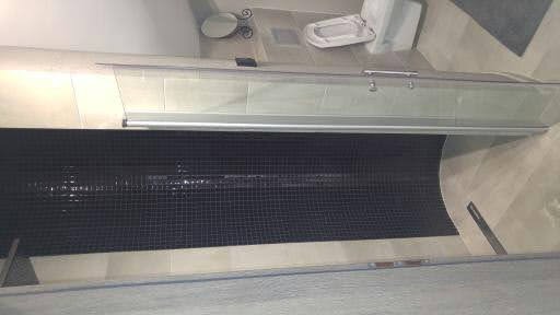 Wykończona łazienka z kabiną prysznicową z przesuwnymi drzwiami, czarną mozaiką na ścianie i jasnymi płytkami podłogowymi, widoczna toaleta i okrągłe lustro.