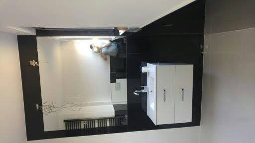 Montaż dużego lustra łazienkowego z czarną ramą, widoczne podłączenie elektryczne, biała szafka z umywalką i baterią, szare płytki na ścianie w tle.