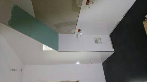 Wnętrze w trakcie prac wykończeniowych, widoczna płyta kartonowo-gipsowa na ścianie, przygotowane instalacje elektryczne i czarna podłoga w remoncie.