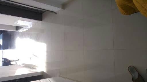 Błyszczące, białe płytki ceramiczne na ścianie łazienki, z odbiciem światła w fugach i widocznym fragmentem sufitu oraz podłogi, z perspektywy pod kątem.