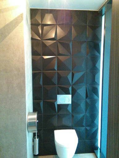 Nowoczesna łazienka z czarnymi, trójwymiarowymi płytkami ściennymi, podwieszaną toaletą i srebrnym uchwytem na papier toaletowy.