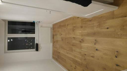 Jasna drewniana podłoga ułożona w nowym mieszkaniu, widok na ścianę z oknem i roletą, fragment sufitu z listwą przypodłogową.