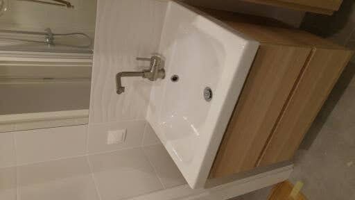 Biała umywalka z drewnianą szafką z trzema szufladami, chromowana bateria w łazience wyłożonej białymi płytkami, widoczne odbicie lustra.