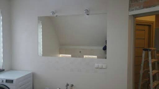 Prostokątne lustro z oświetleniem nad umywalką w łazience z białymi płytkami, widoczny fragment pralki i drabina w tle.