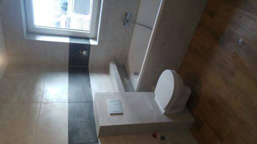 Wykończona łazienka z beżowymi i czarnymi płytkami, drewnianą podłogą, białą wanną i podwieszanym WC z przyciskiem spłukującym.