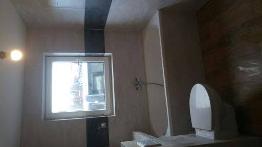 Wykończona łazienka z wanną, toaletą i oknem, z widocznymi płytkami w odcieniach beżu i czarnymi akcentami, oświetlona pojedynczą żarówką.