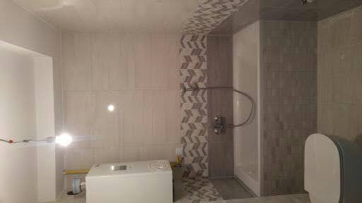 Wnętrze łazienki w trakcie remontu, widoczne płytki na ścianach, kabina prysznicowa i częściowo zamontowana toaleta, na podłodze i ścianach wzór w jodełkę, w rogu pomieszczenia stoi biały zbiornik.