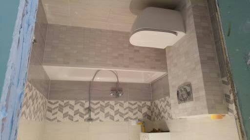 Wykończona łazienka z szaro-białymi płytkami w geometryczny wzór na ścianach, widoczna wanna z baterią i podwieszana toaleta z zabudowaną spłuczką.