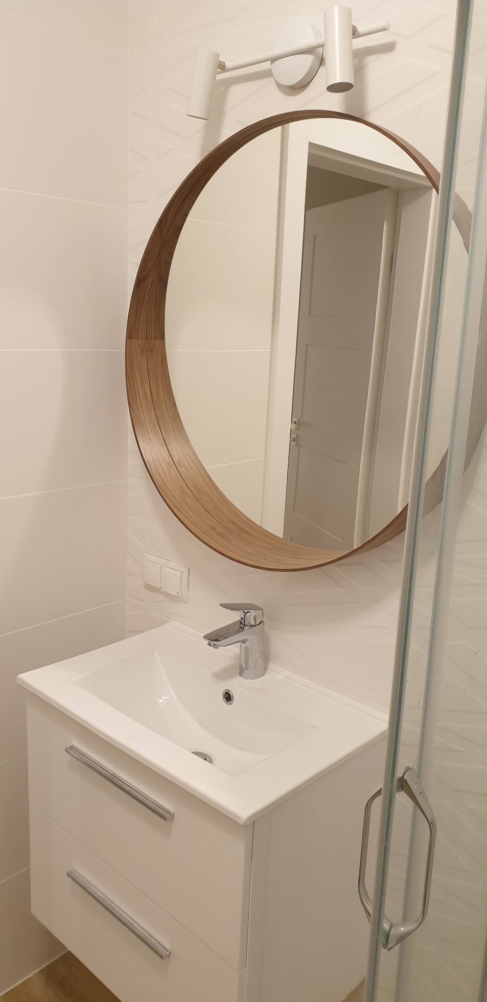 Nowoczesna łazienka z białą umywalką nablatową na szafce z dwiema szufladami, okrągłym lustrem w drewnianej ramie i subtelnym oświetleniem punktowym.