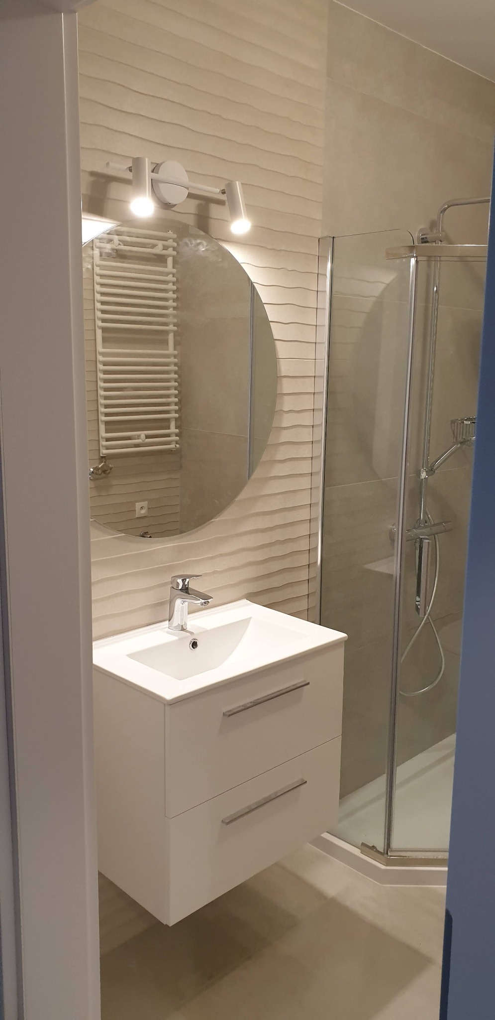 Nowoczesna łazienka z umywalką na szafce z dwoma szufladami, okrągłym lustrem z oświetleniem i kabiną prysznicową w tle, ściany wykończone płytkami z delikatnym wzorem fal.