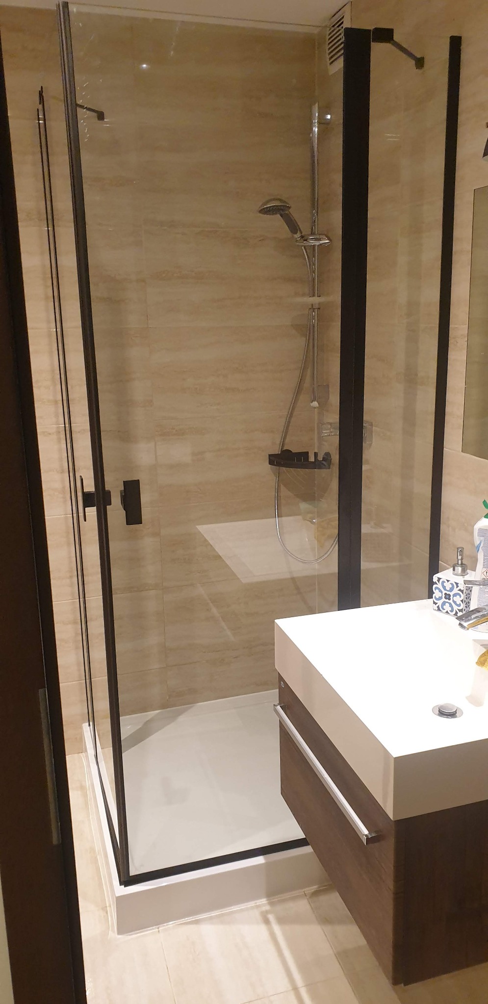 Nowoczesna kabina prysznicowa z czarną ramą i deszczownicą, beżowe płytki na ścianach, biała umywalka z szafką w kolorze drewna.