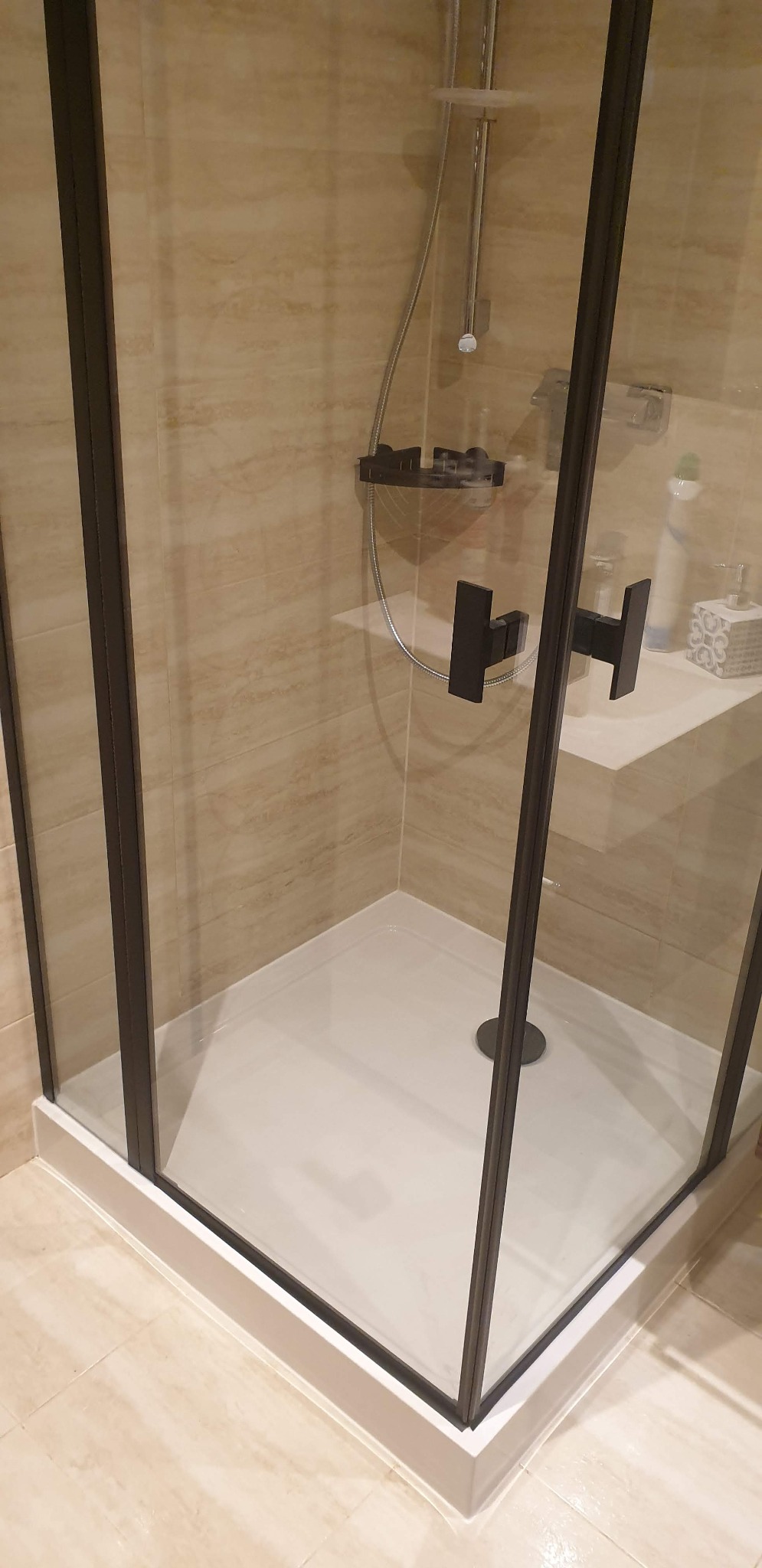 Nowoczesna kabina prysznicowa z czarnymi profilami, brodzikiem w kolorze białym i beżowymi płytkami imitującymi marmur na ścianach, z widoczną półką na kosmetyki i czarnym uchwytem na słuchawkę...