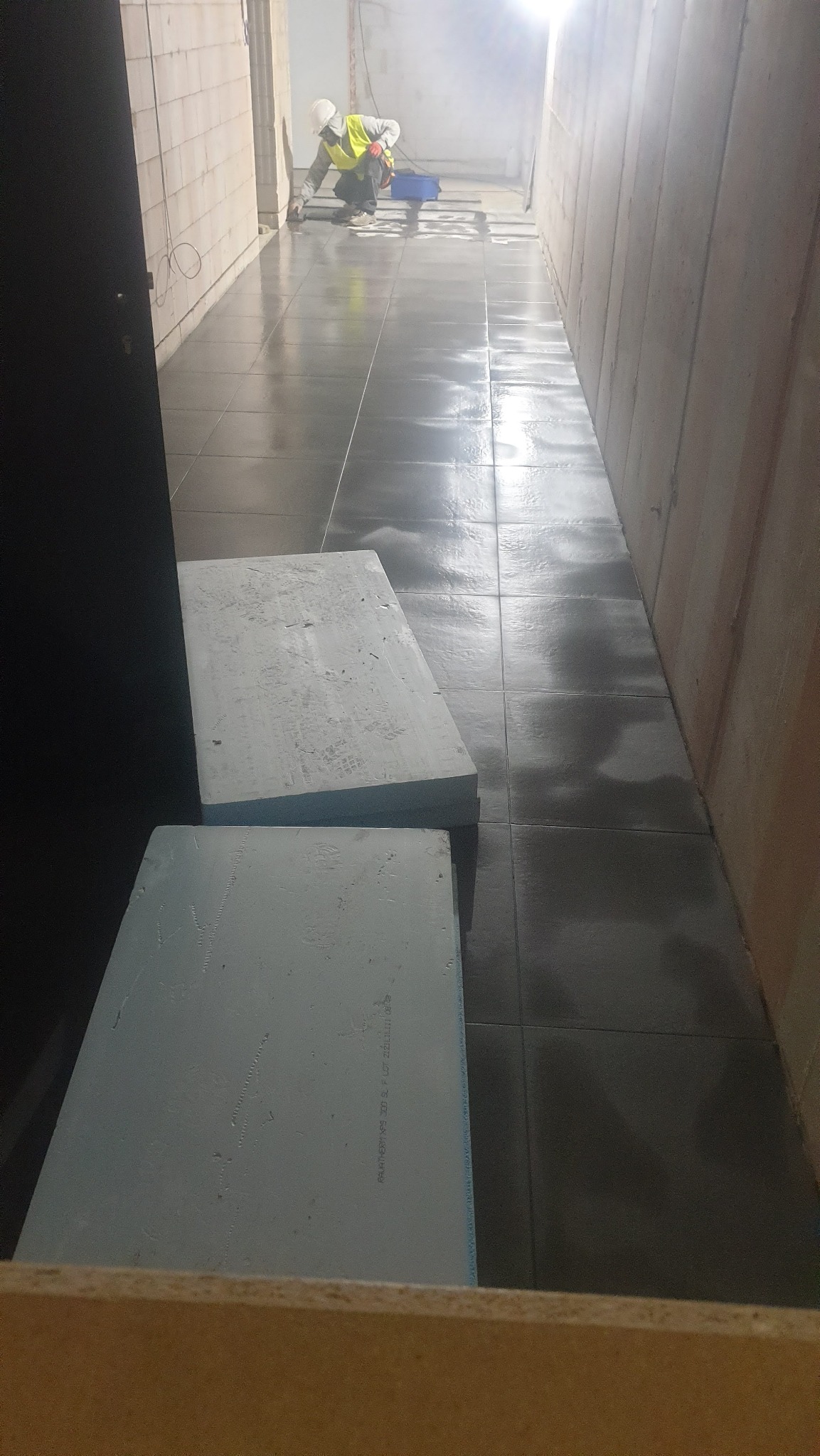 Korytarz w trakcie układania ciemnych płytek podłogowych, pracownik w odblaskowej kamizelce fuguje spoiny, w kadrze widoczne płyty styropianowe.