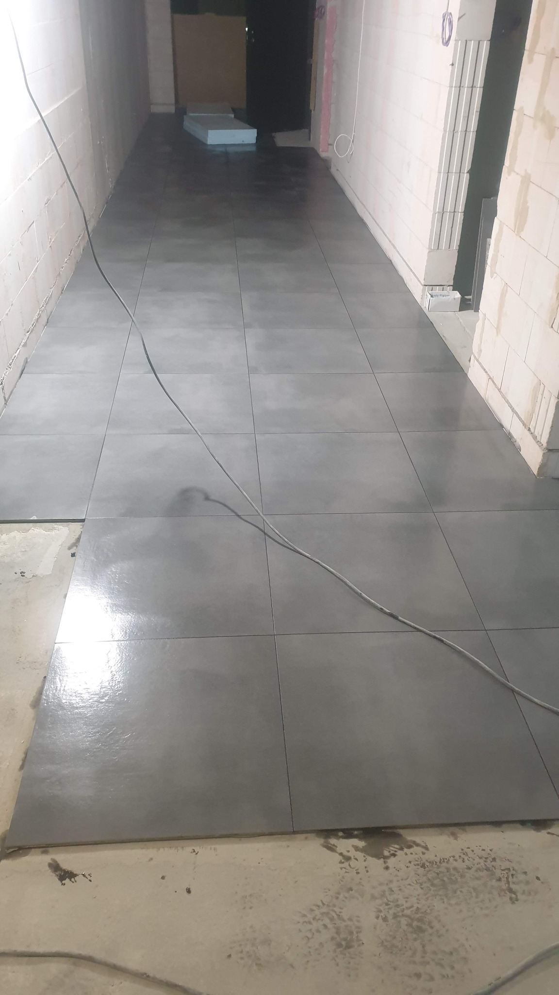 Układanie szarych, matowych płytek podłogowych w długim, wąskim pomieszczeniu; widoczne niedociągnięcia przy ścianie i przewód elektryczny na podłodze.