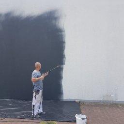 Intermal - Mężczyzna w odzieży roboczej maluje ścianę zewnętrzną natryskowo na szaro, widoczny fragment pomalowanej i niepomalowanej powierzchni, wiadro z farbą i zabezpieczająca folia na ziemi.