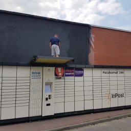 Intermal - Mężczyzna maluje ciemną farbą elewację budynku nad Paczkomatem InPost, widoczne zabezpieczenia folią malarską.