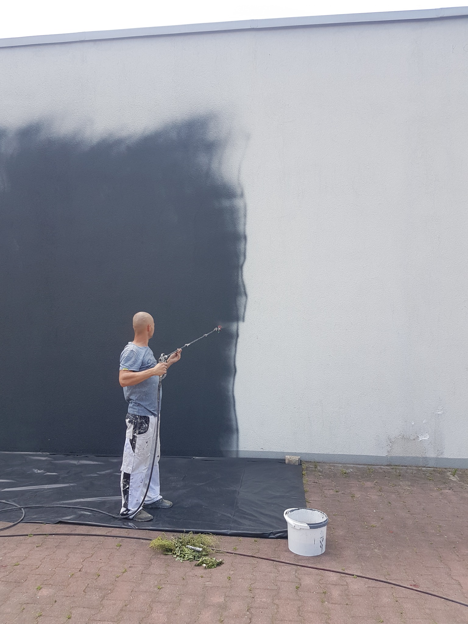 Mężczyzna w odzieży roboczej maluje ścianę zewnętrzną natryskowo na szaro, widoczny fragment pomalowanej i niepomalowanej powierzchni, wiadro z farbą i zabezpieczająca folia na ziemi.