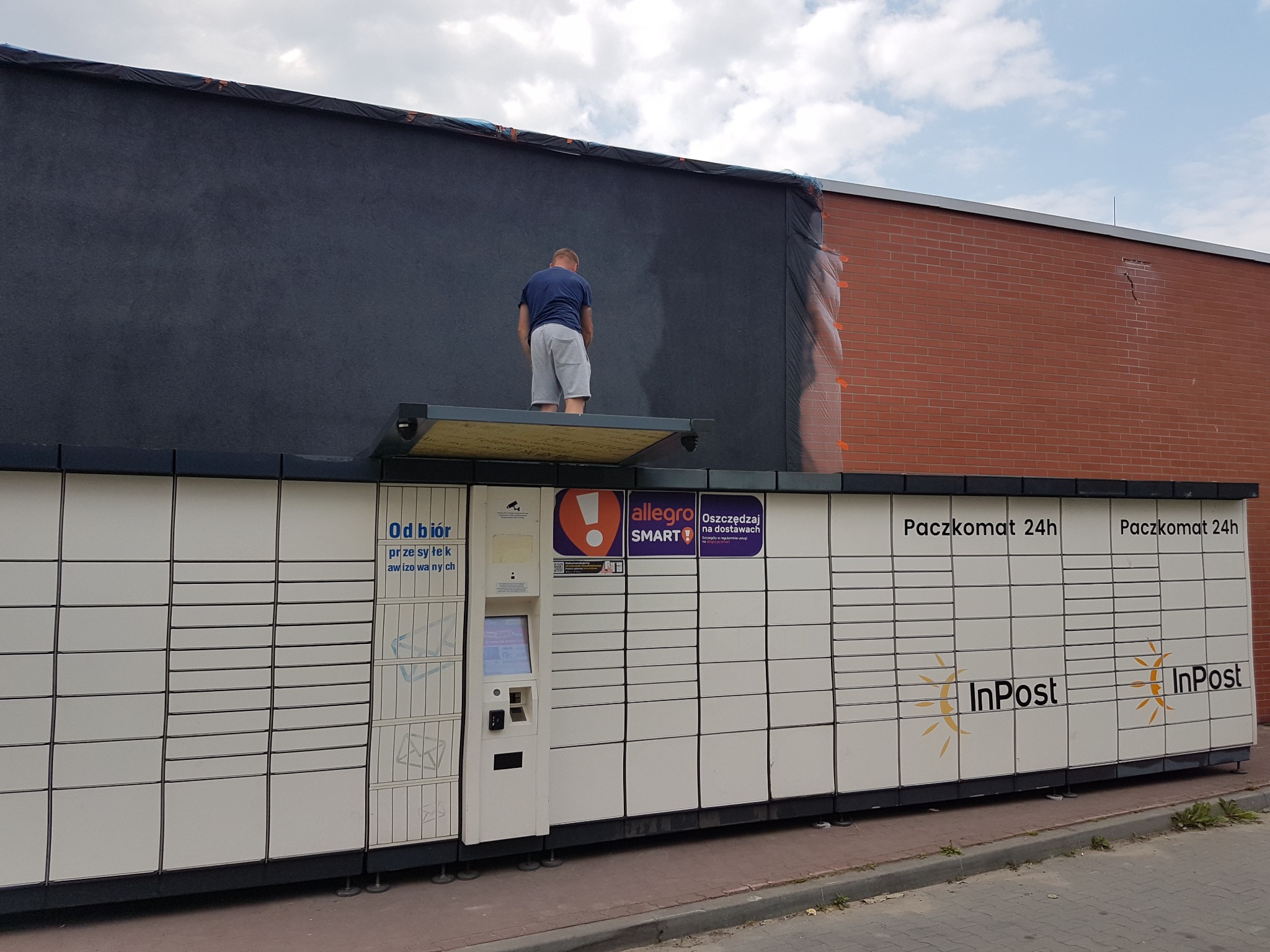 Mężczyzna maluje ciemną farbą elewację budynku nad Paczkomatem InPost, widoczne zabezpieczenia folią malarską.