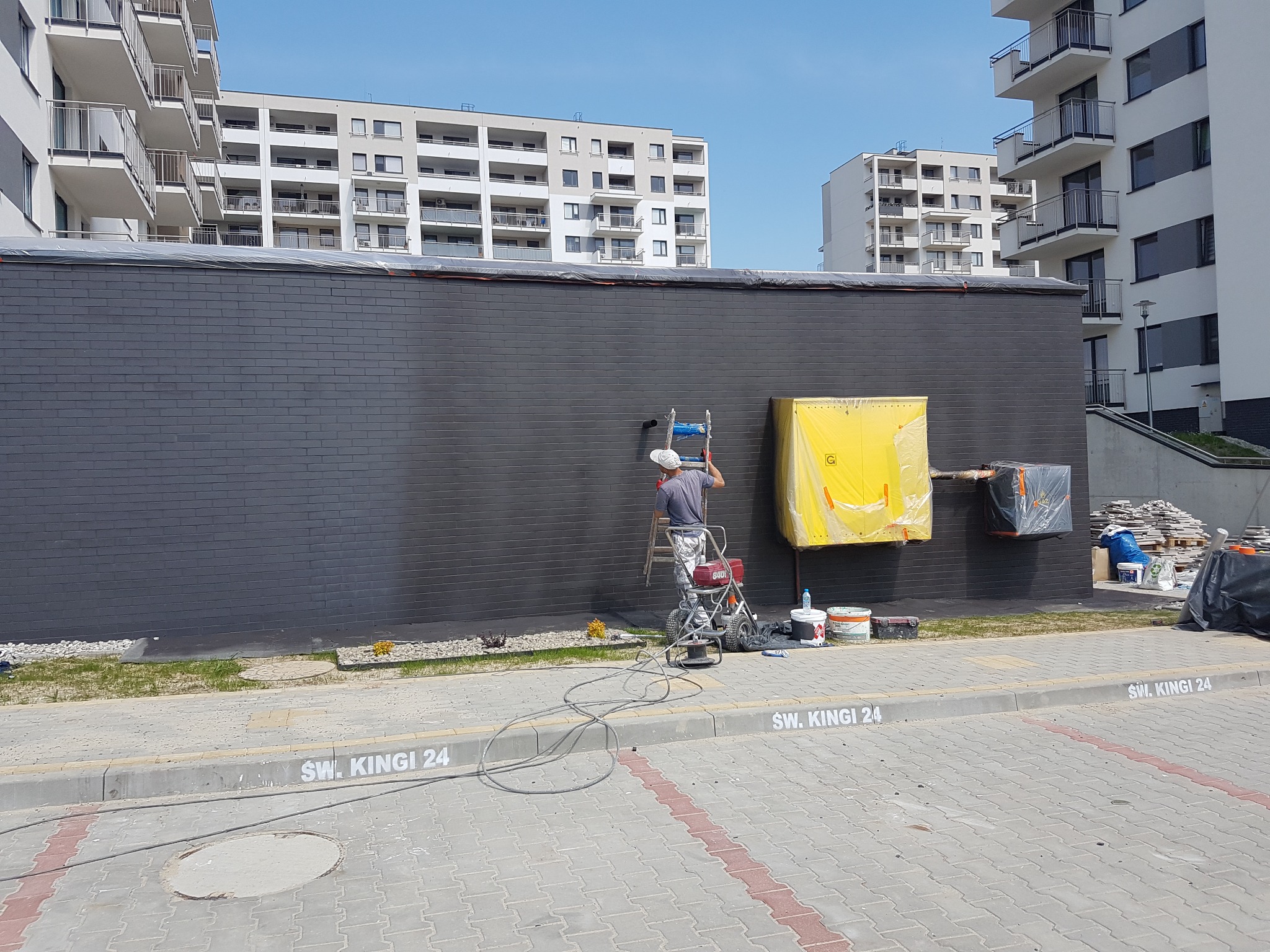 Mężczyzna na drabinie maluje ciemnoszarą ścianę budynku z cegły, używając pistoletu natryskowego; w tle widoczne bloki mieszkalne i chodnik z napisem 'Św. Kingi 24'.