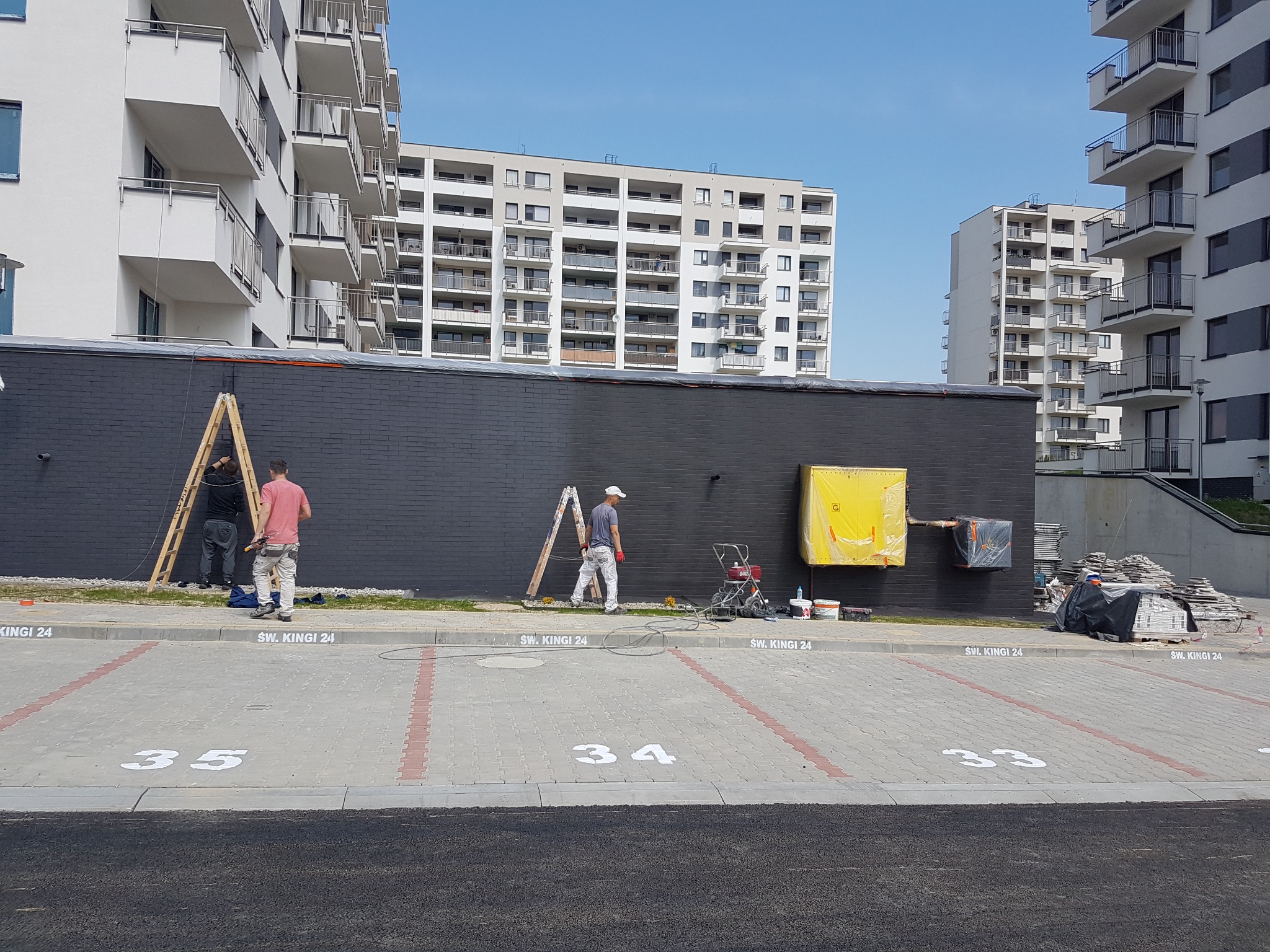 Dwóch pracowników maluje ciemną ścianę budynku mieszkalnego, jeden na drabinie, drugi z agregatem malarskim na parkingu z numerami miejsc.