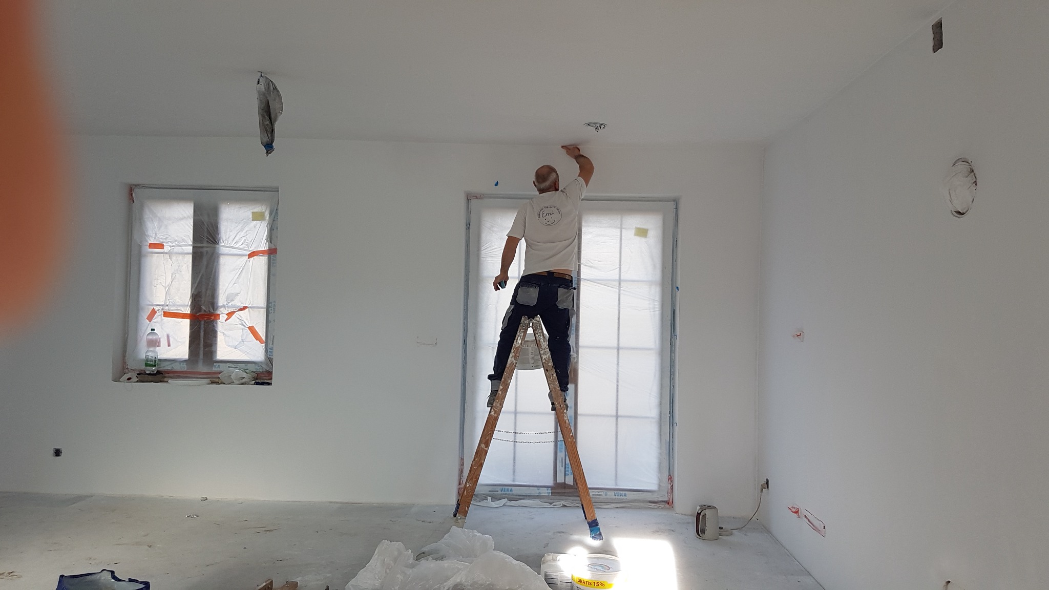 Mężczyzna na drabinie maluje sufit w pustym, białym pokoju. Okna i drzwi zabezpieczone folią malarską. Widoczne puszki z farbą i worki foliowe na podłodze.