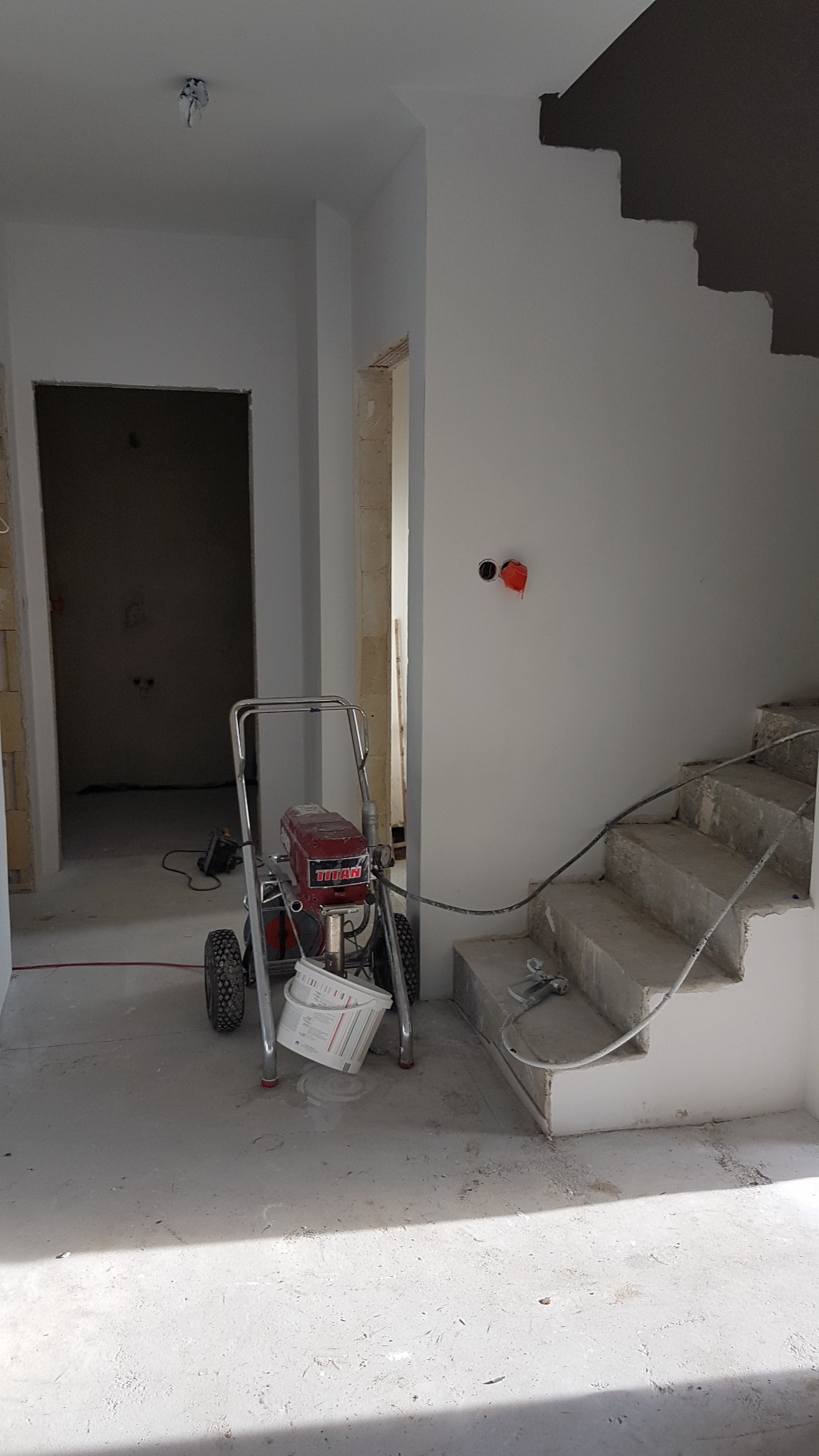 Wnętrze w trakcie prac wykończeniowych: widoczny agregat malarski Titan na tle surowych schodów i świeżo pomalowanych ścian na biało; w tle otwarte przejście do kolejnego pomieszczenia.