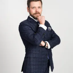 Portret mężczyzny w granatowym garniturze w kratę, z ręką podpierającą brodę, na jasnym tle.