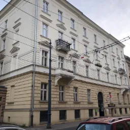 Odnowiona, kremowo-biała fasada kamienicy z balkonami, widok z poziomu ulicy, z widocznymi samochodami i słupem oświetleniowym.