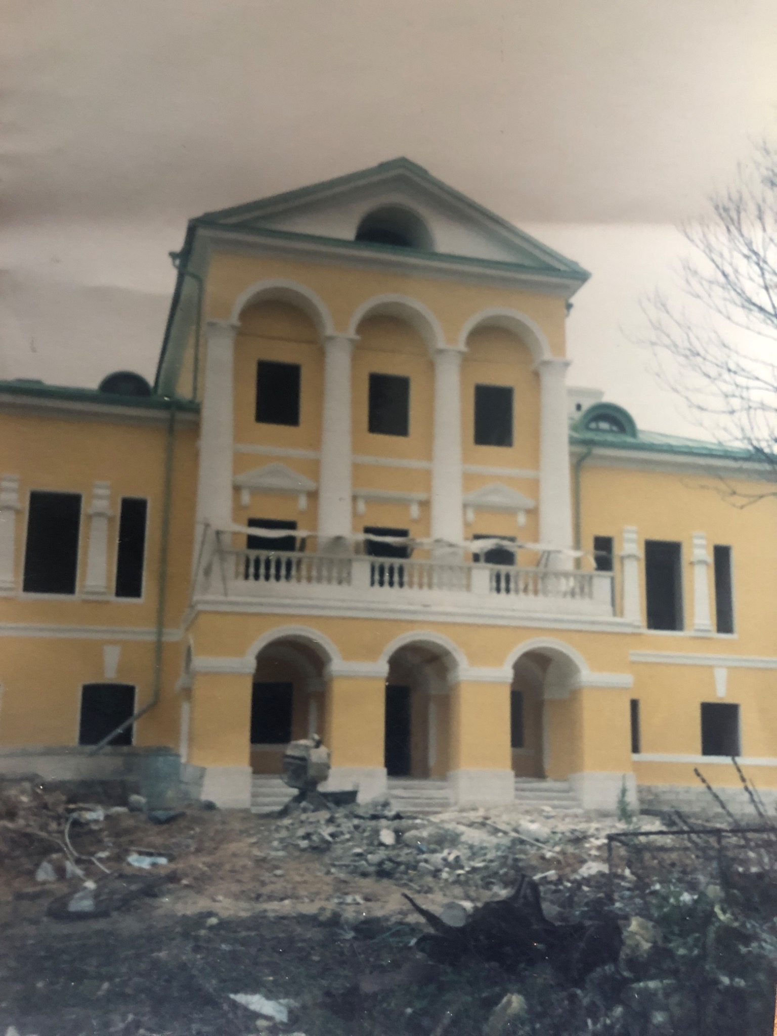 Żółty budynek z białymi kolumnami i łukami, prawdopodobnie w trakcie prac renowacyjnych; na pierwszym planie gruz i pozostałości po budowie, drzewo bez liści po prawej stronie.