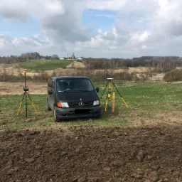 Czarny van Mercedes i dwa geodezyjne odbiorniki GPS na trójnogach, ustawione na polu z przygotowaną ziemią, w tle pagórkowaty krajobraz i pochmurne niebo.