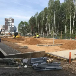 Przygotowanie terenu pod budowę drogi: walec drogowy ubija ziemię, w tle koparka i wywrotka, widoczne pryzmy ziemi, krawężniki i elementy konstrukcyjne.