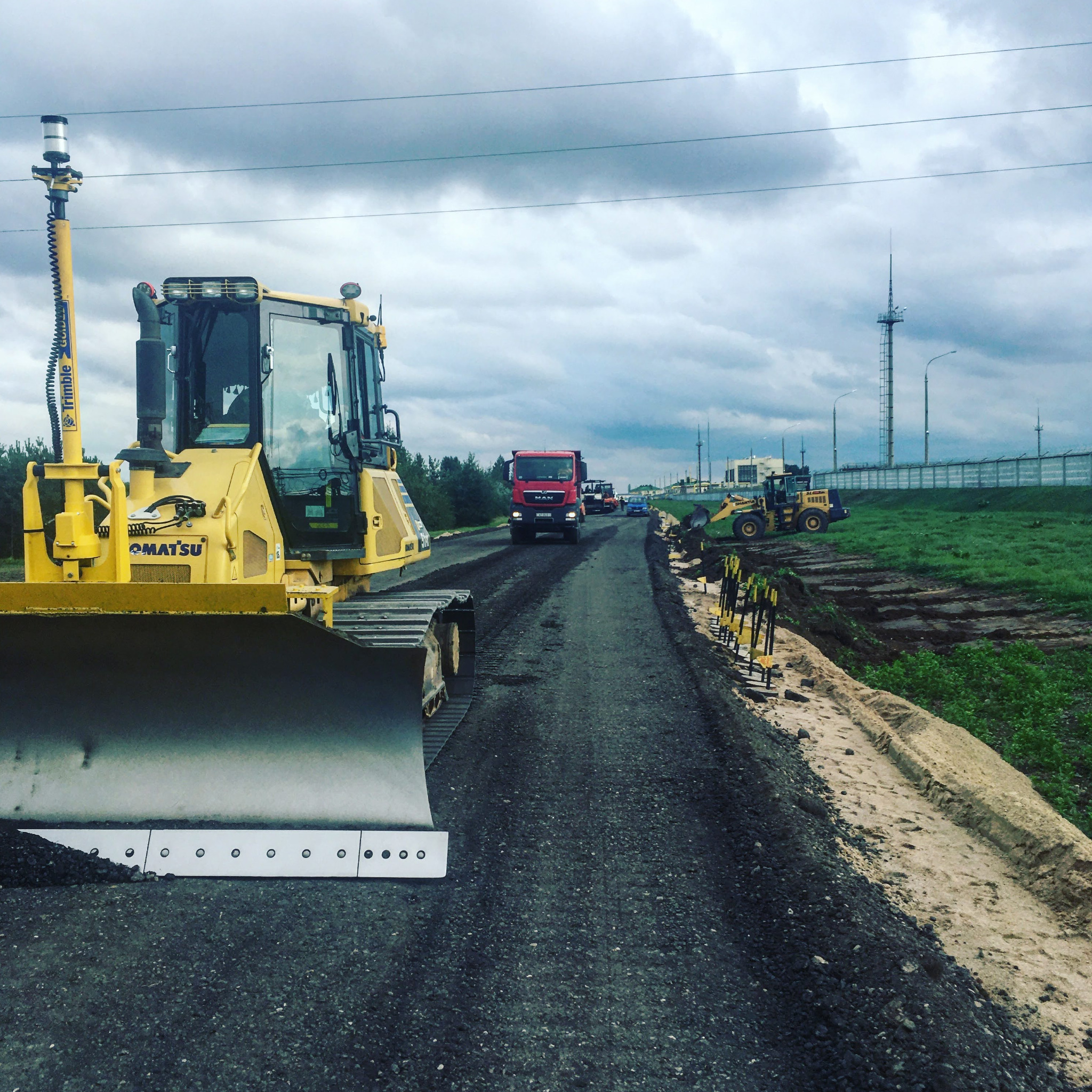 Żółty buldożer Komatsu z systemem GPS Trimble wyrównuje świeżo położony asfalt na budowie drogi. W tle widoczny czerwony samochód ciężarowy MAN i inne maszyny budowlane oraz pochmurne niebo.