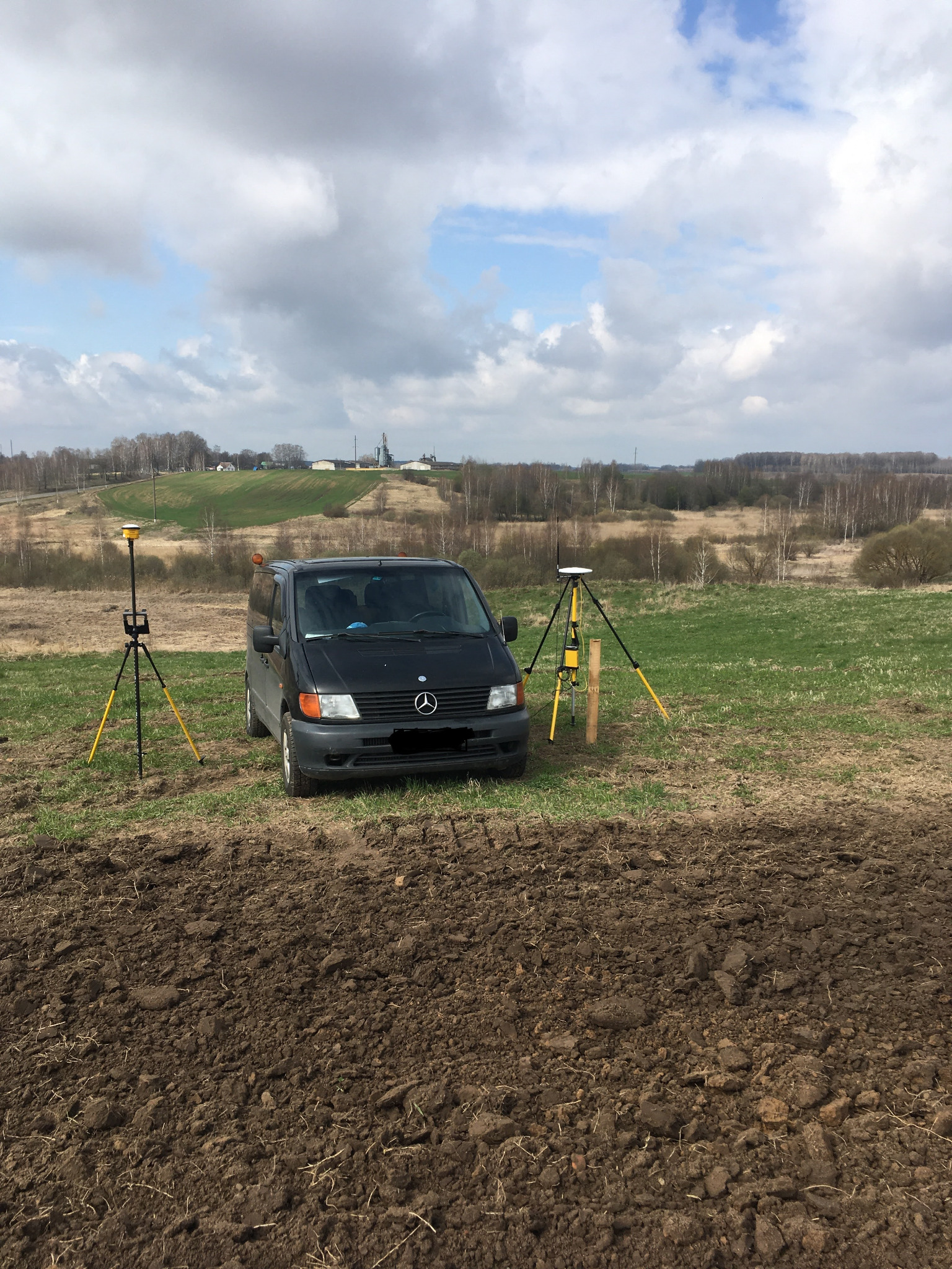 Czarny van Mercedes i dwa geodezyjne odbiorniki GPS na trójnogach, ustawione na polu z przygotowaną ziemią, w tle pagórkowaty krajobraz i pochmurne niebo.