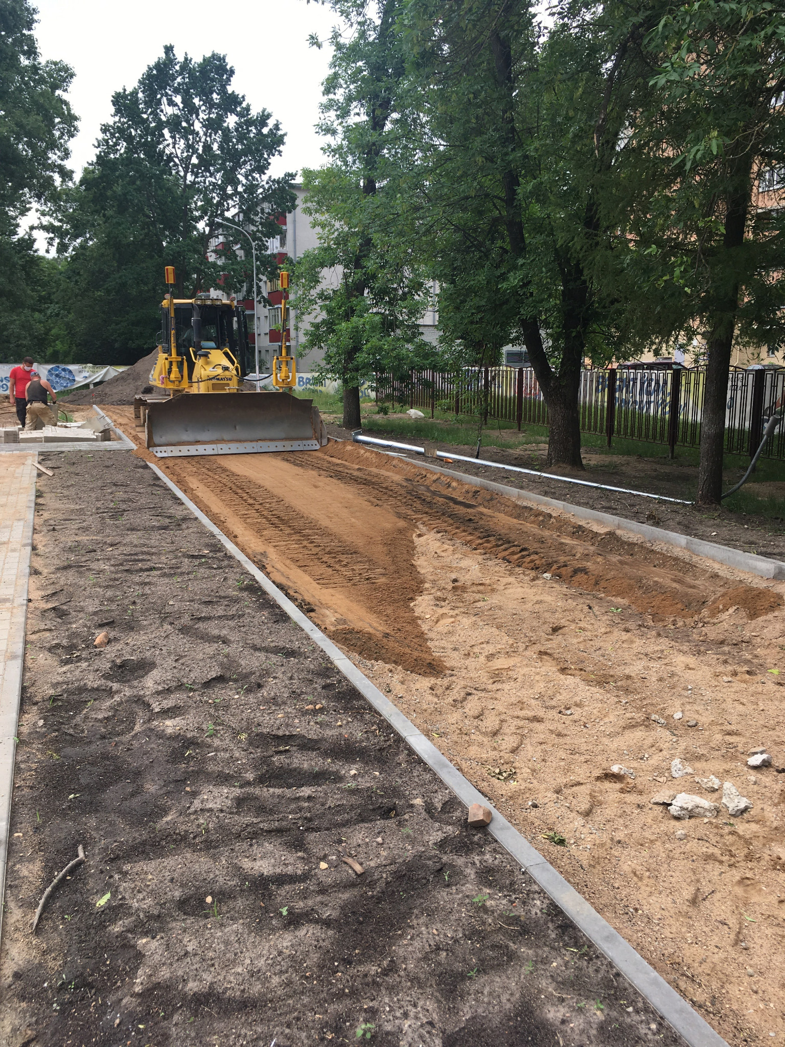 Żółty spychacz wyrównuje teren pomiędzy betonowymi krawężnikami, przygotowując podłoże pod budowę chodnika lub drogi, z widocznymi śladami gąsienic na piasku.