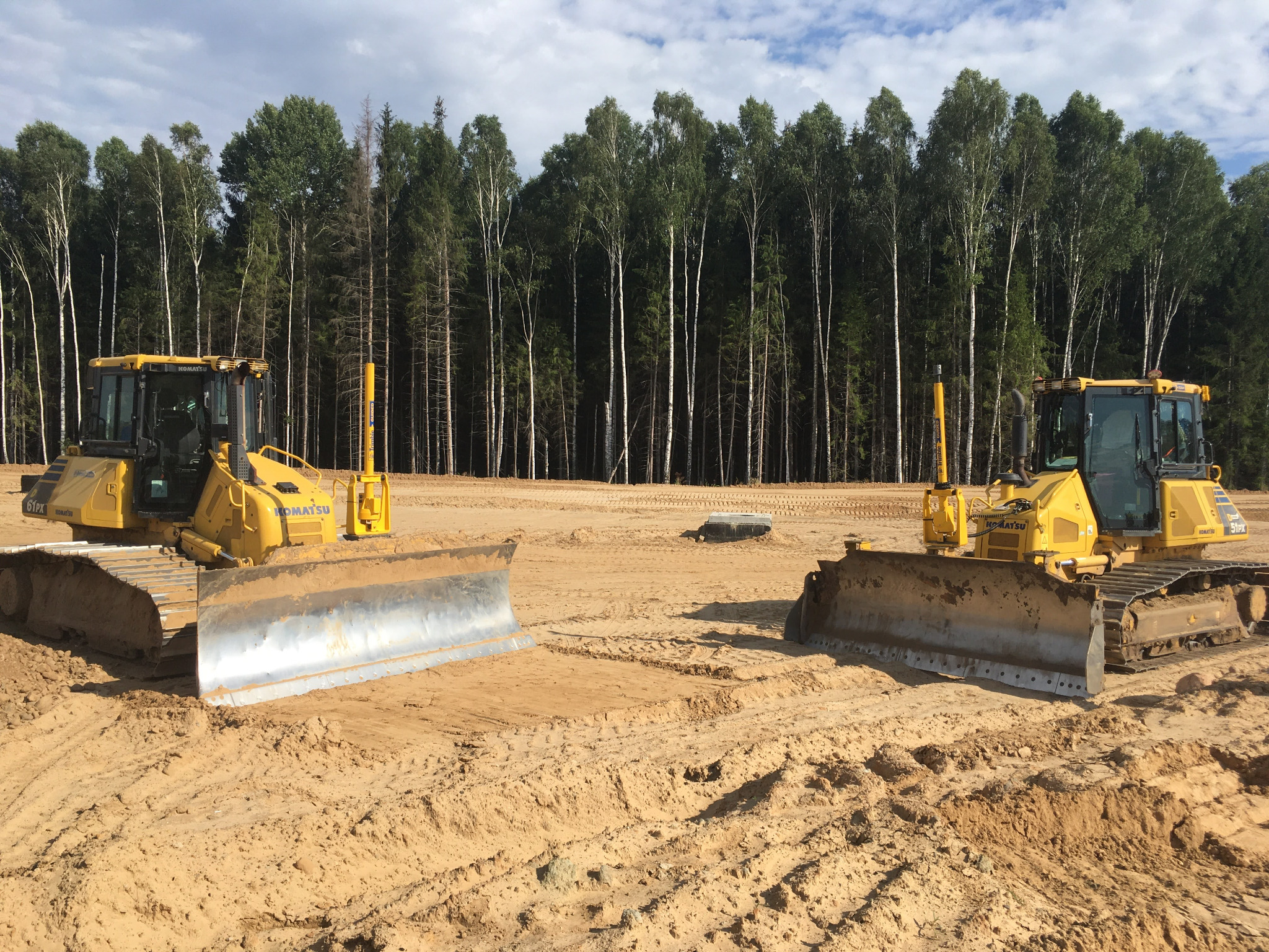 Dwa żółte spychacze Komatsu, ustawione naprzeciwko siebie na wyrównanym, piaszczystym terenie z widocznymi śladami gąsienic, w tle wysoki las liściasty pod błękitnym niebem z chmurami.