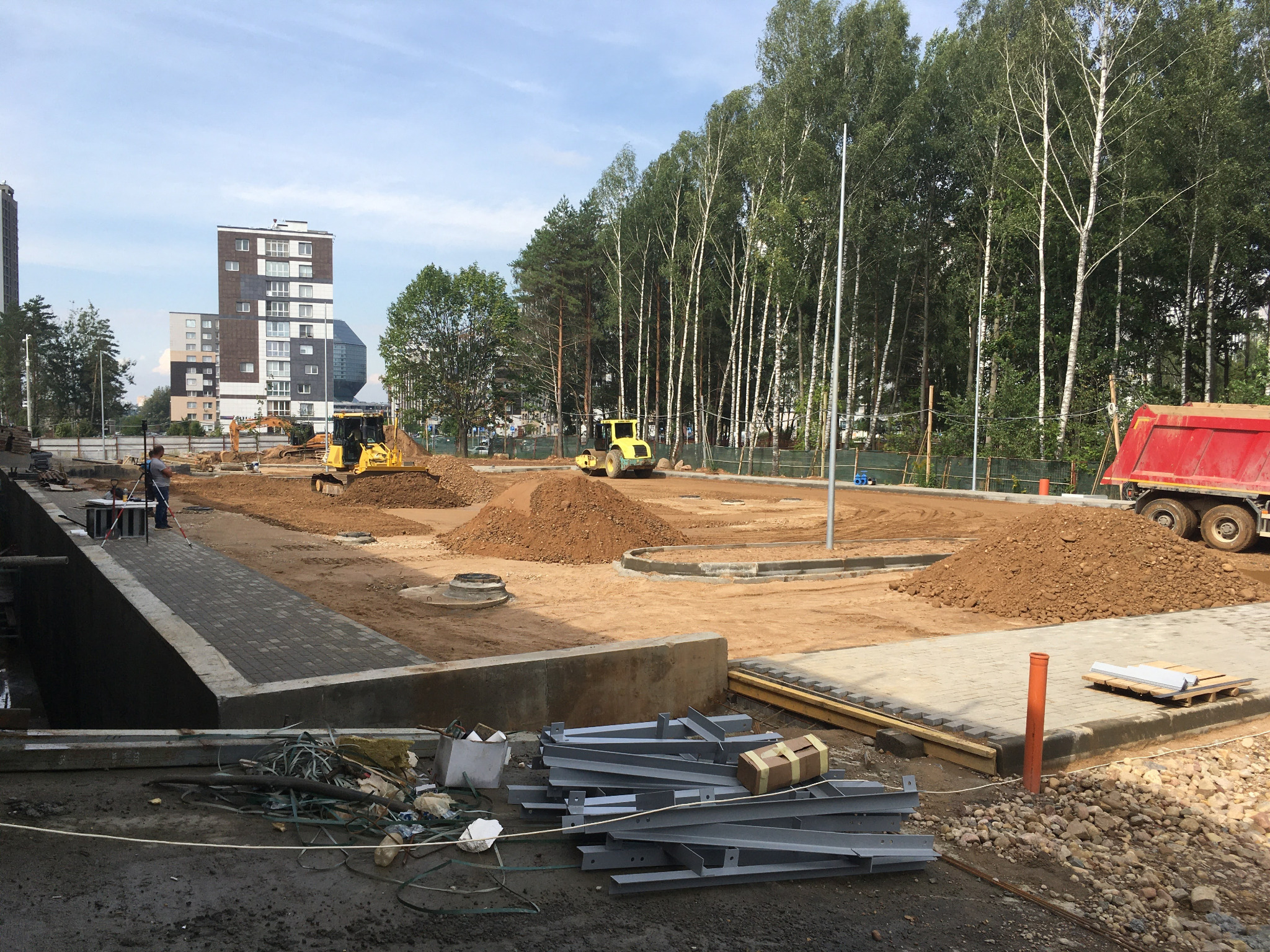 Przygotowanie terenu pod budowę drogi: walec drogowy ubija ziemię, w tle koparka i wywrotka, widoczne pryzmy ziemi, krawężniki i elementy konstrukcyjne.