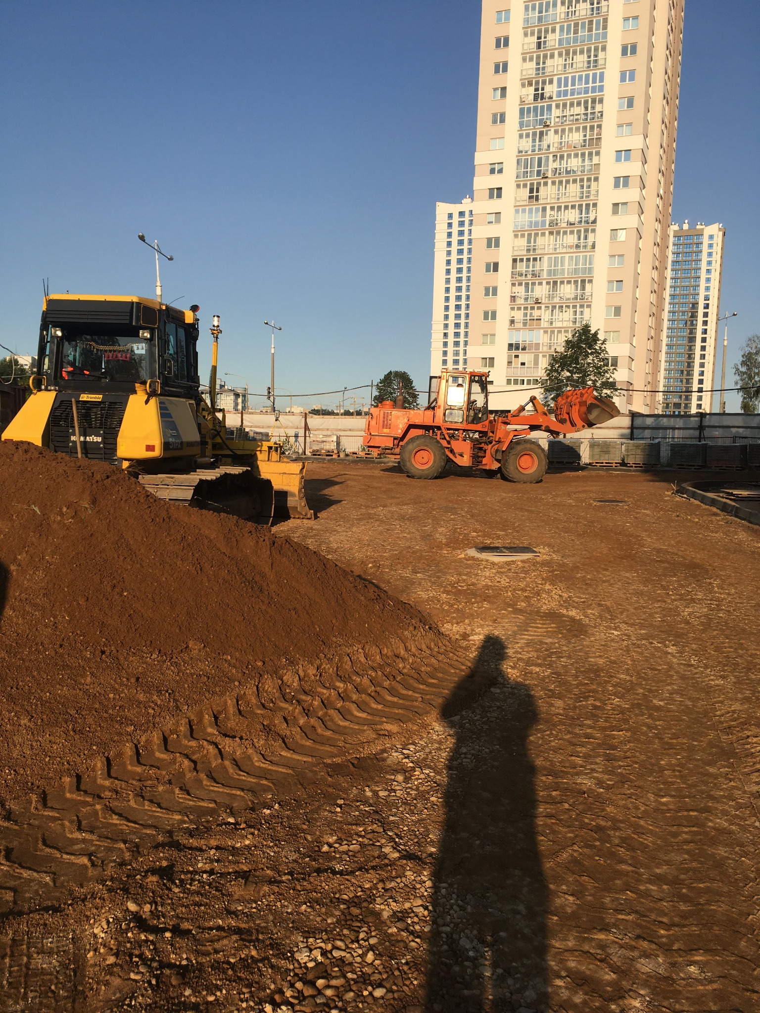 Żółta spycharka Komatsu i pomarańczowa ładowarka kołowa na placu budowy z pryzmą ziemi w pierwszym planie, w tle wysokie bloki mieszkalne i cień osoby.